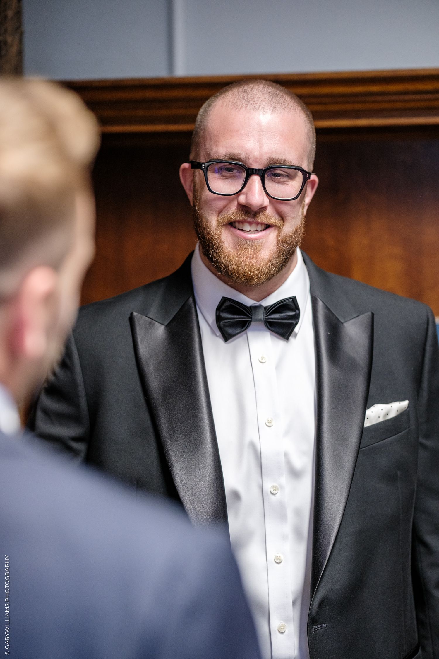 Smiling groom in black tuxedo and bow tie with glasses and red beard at wedding ceremony.