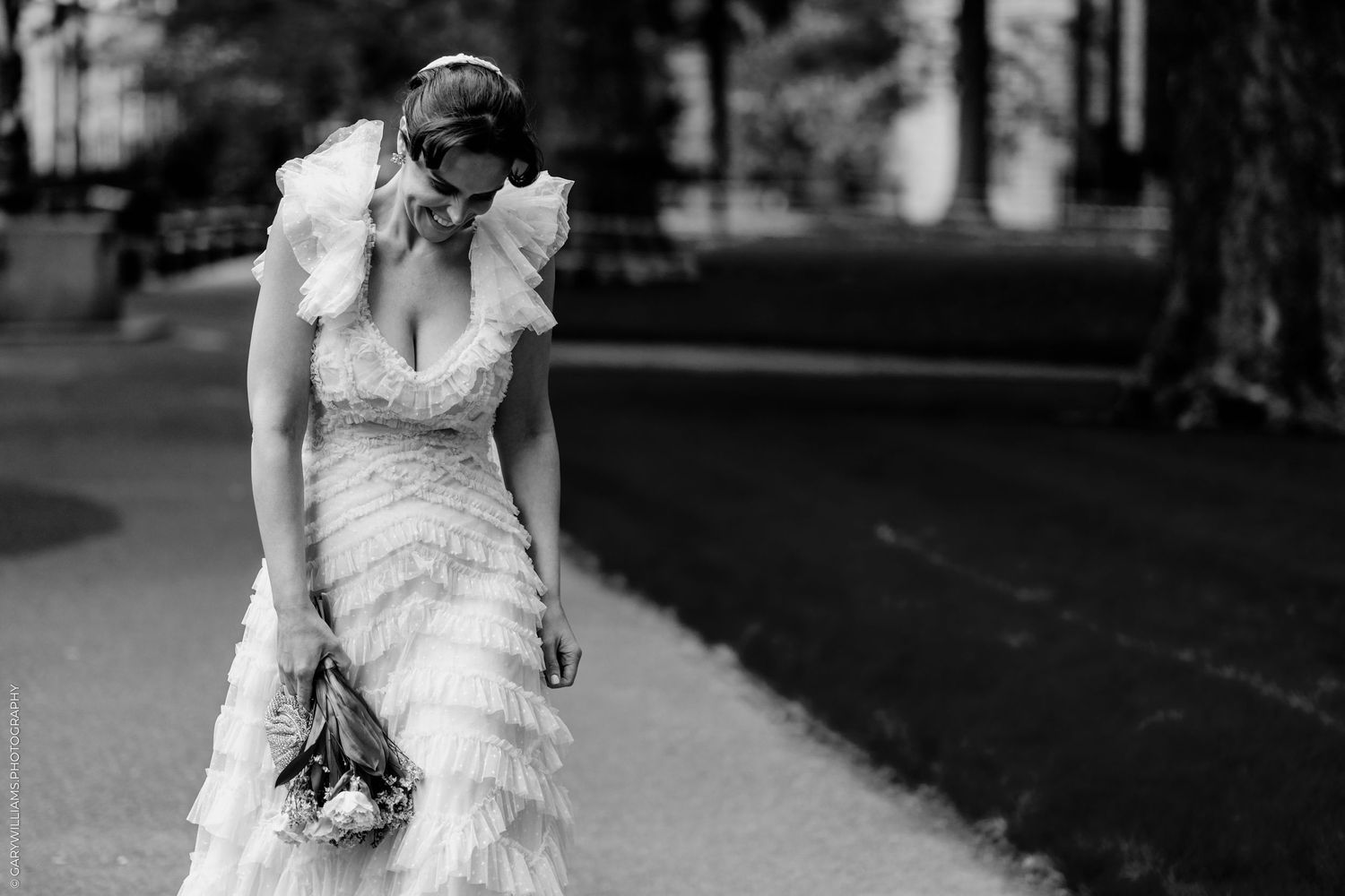 Black and white photo of a bride in a ruffled wedding gown holding a bouquet while looking down in a park.