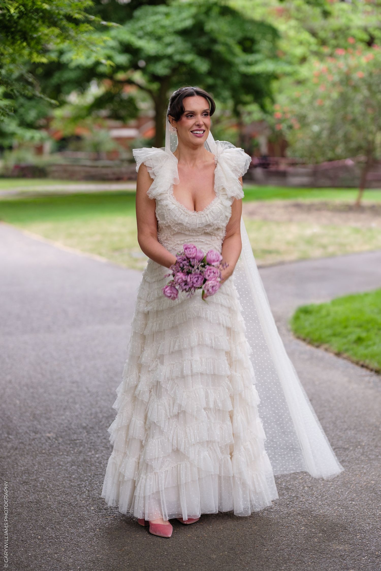 Bride in ruffled white tulle wedding gown with pink bouquet and veil walks along garden path wearing pink heels.