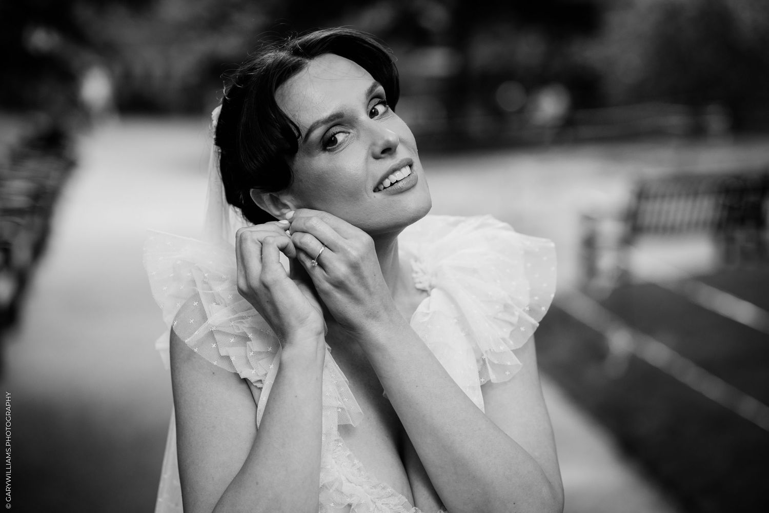 Black and white portrait of a bride adjusting her veil outdoors, wearing a white dress with ruffled collar.