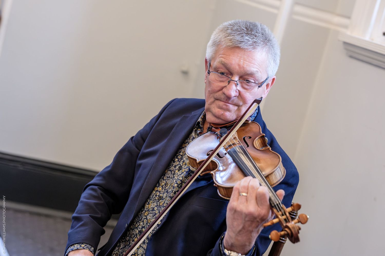 Elderly man playing violin wearing blue blazer and patterned shirt, performing at an indoor music event.