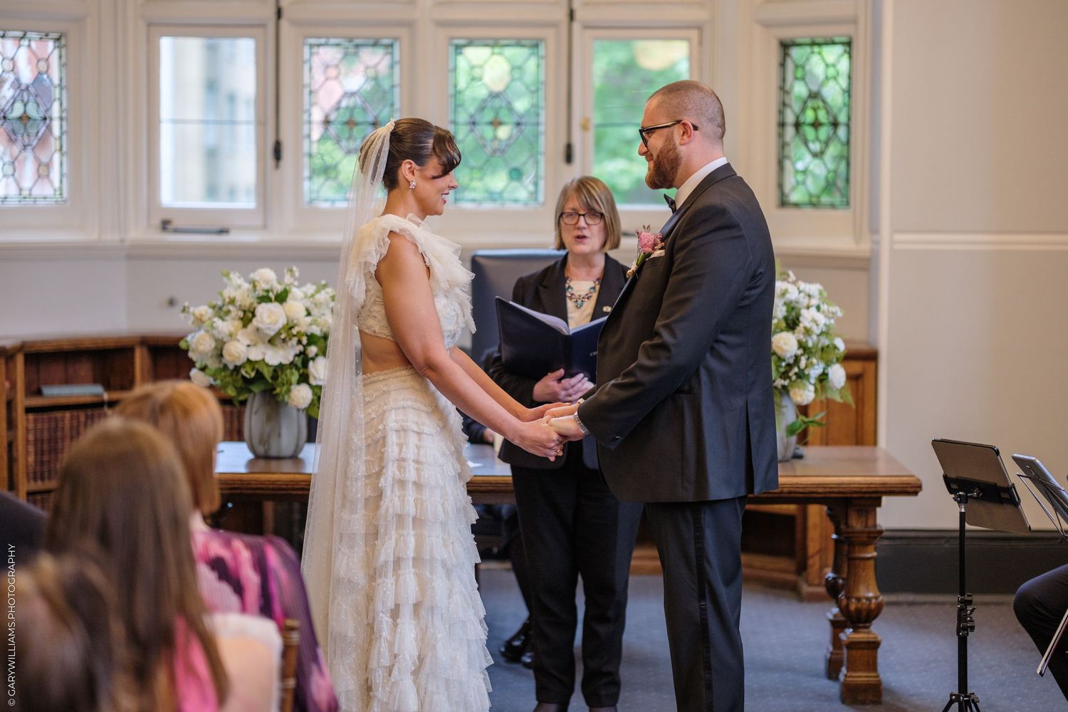 Bride and groom exchange vows at elegant indoor wedding ceremony with officiant and white floral arrangements.