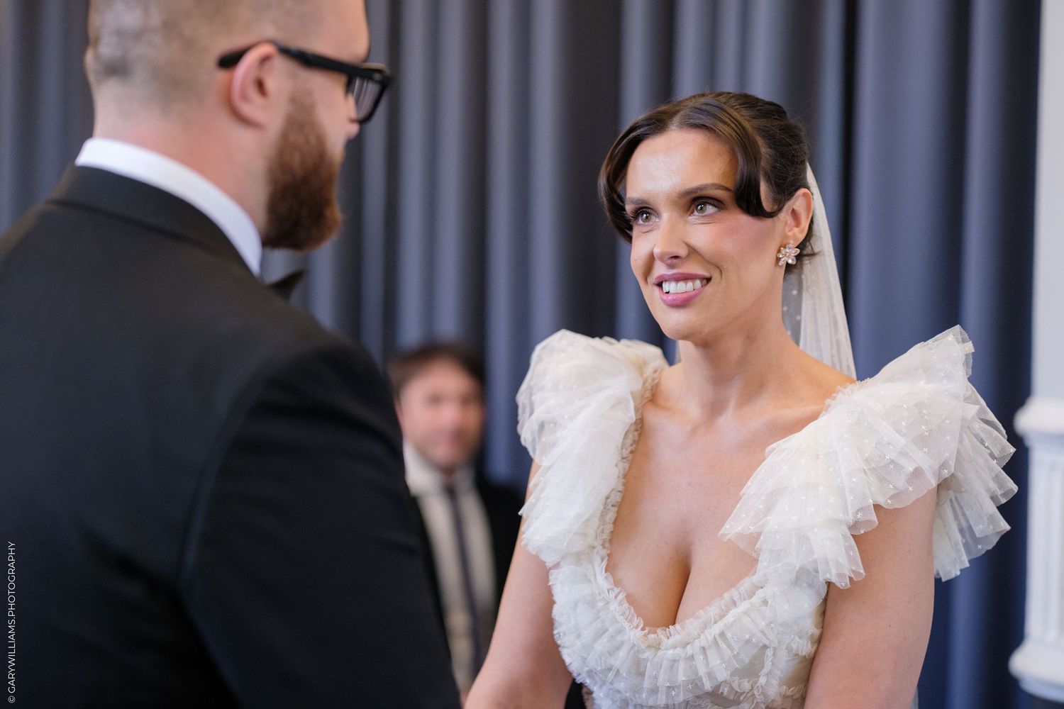 Bride in ruffled white wedding gown smiling at groom during intimate wedding ceremony indoors.