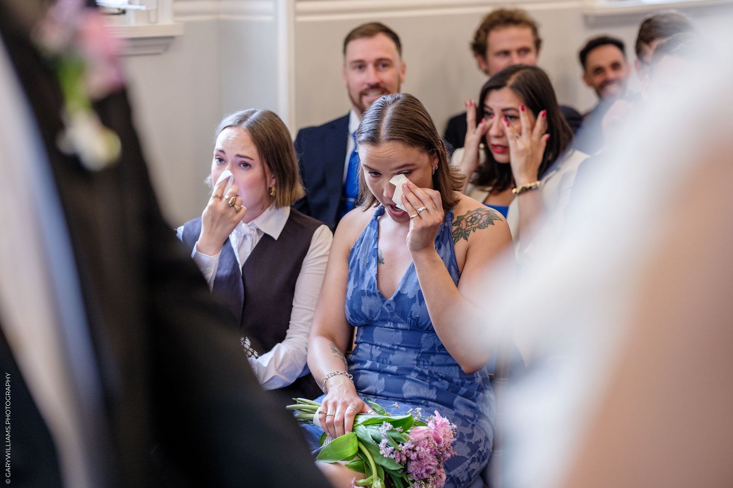 Emotional wedding guests wiping tears during a heartfelt ceremony, with women in blue and grey dresses crying in the front row.