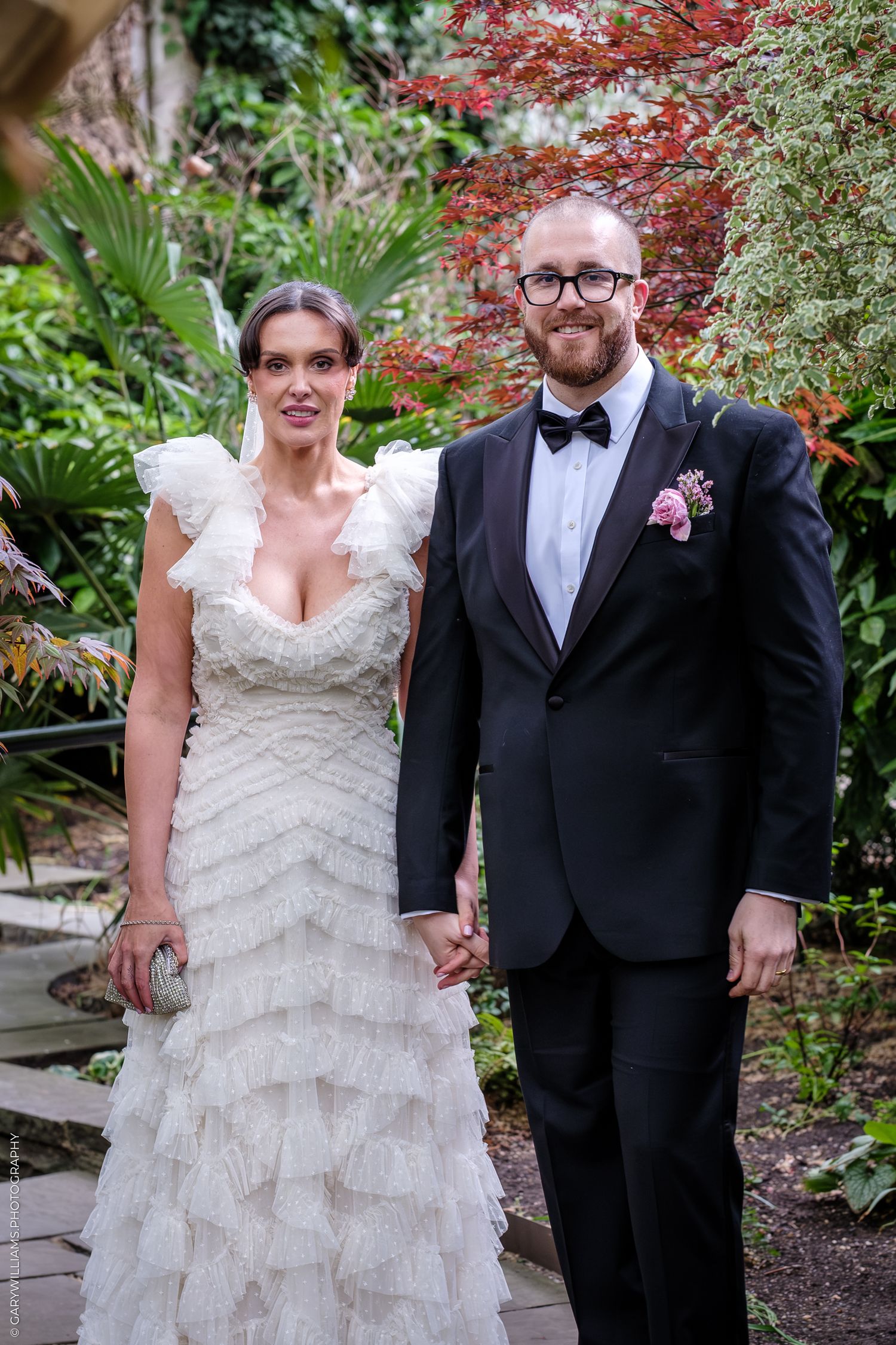 Bride in ruffled white gown and groom in black tuxedo holding hands in a lush garden setting.