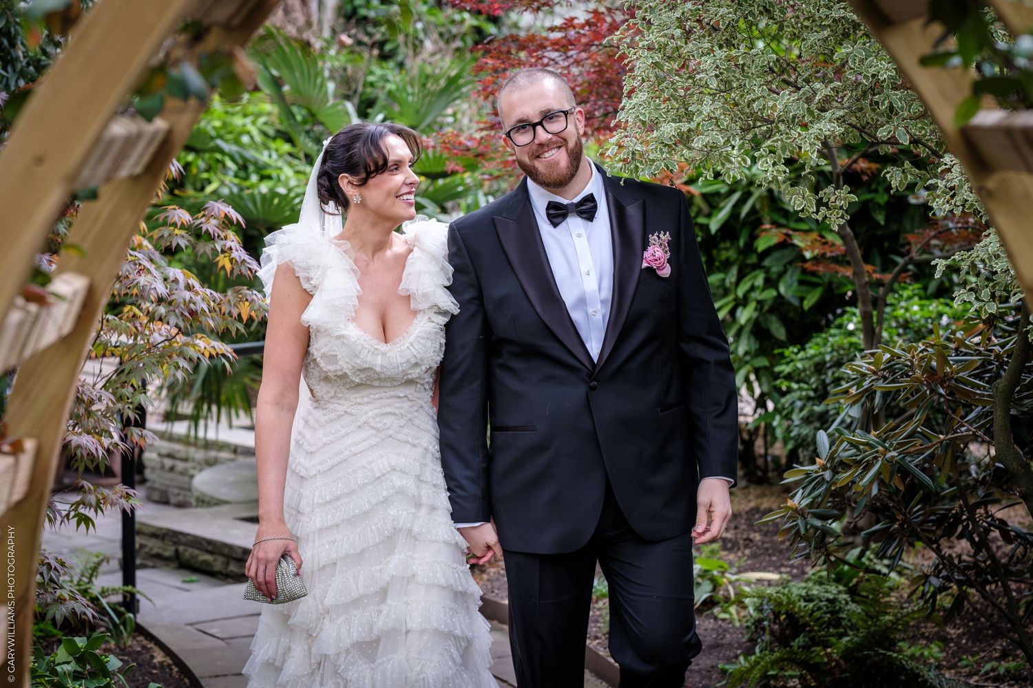 Bride and groom holding hands smiling under a wooden arch surrounded by lush green garden foliage.