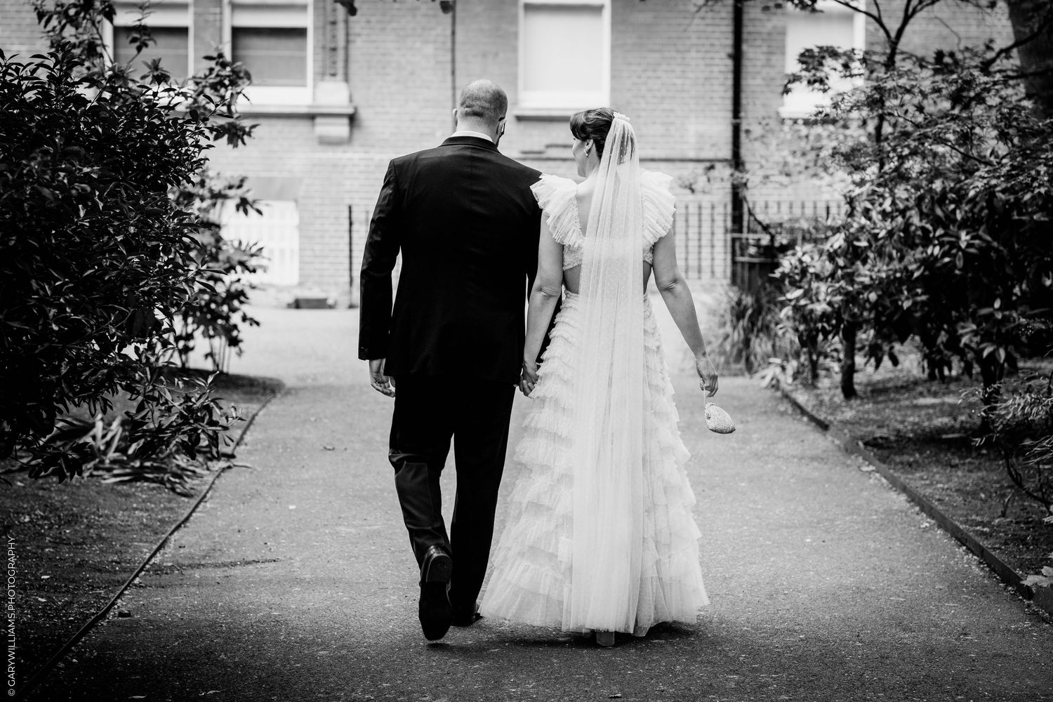 Bride and groom walk hand in hand down a garden path in classic black and white wedding photo.