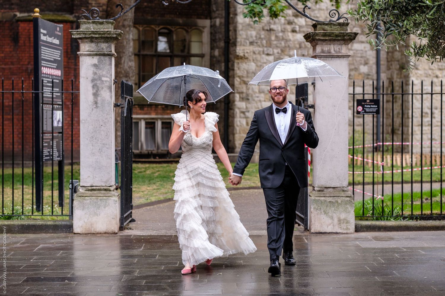 Bride and groom walk hand-in-hand in the rain holding clear umbrellas outside historic stone gates on their wedding day.