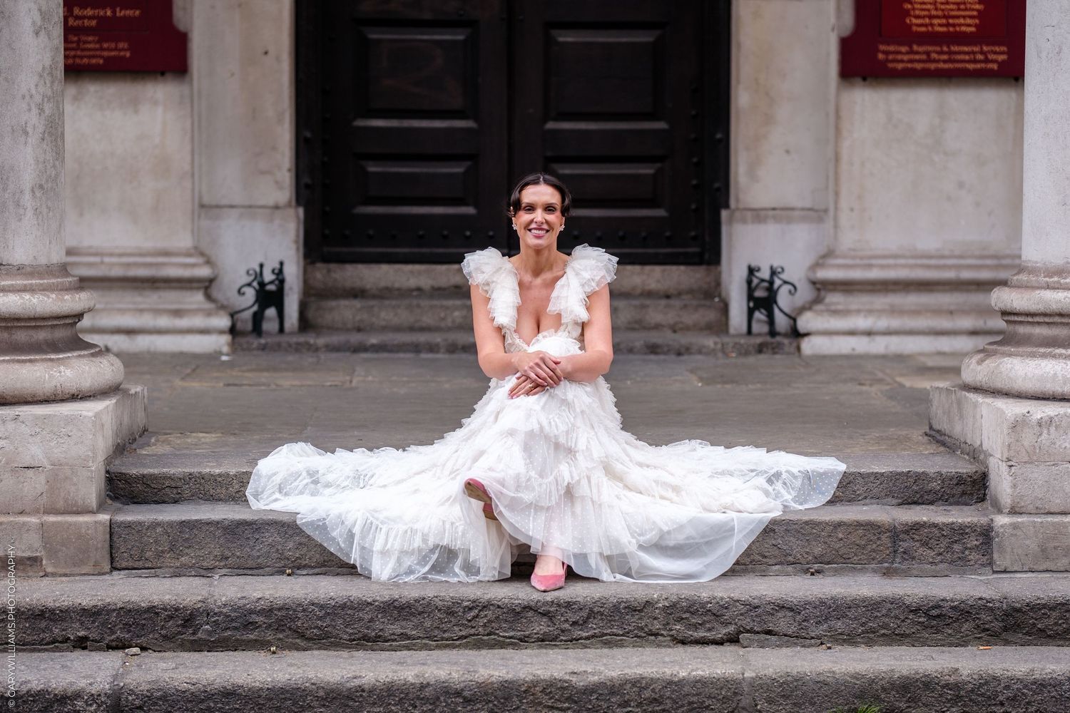 Bride in white gown sitting on stone steps of classical building, smiling joyfully on her wedding day.