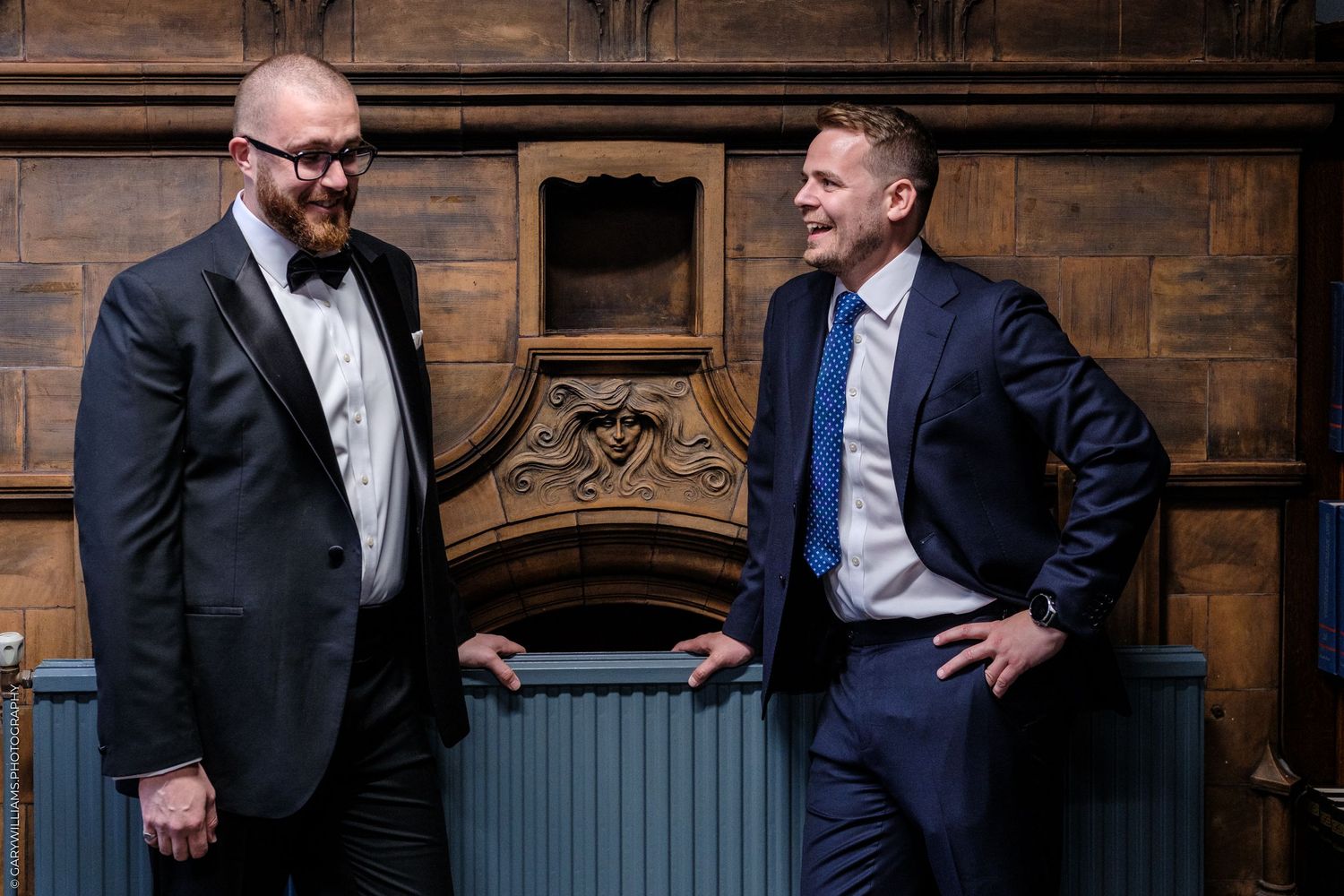 Two men in formal suits smiling at each other near an ornate wooden fireplace mantel.