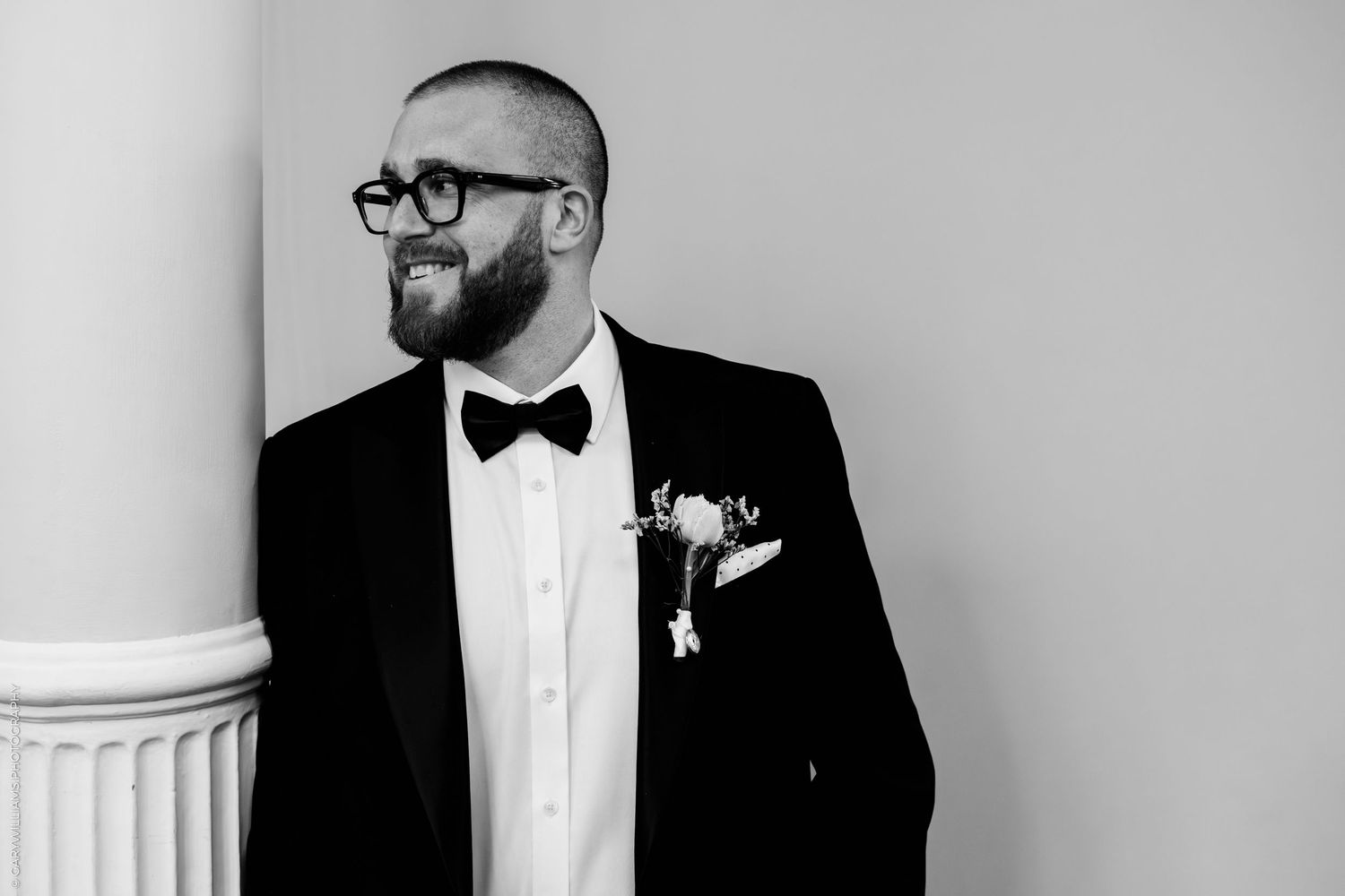 Dapper groom in black tuxedo and bow tie with boutonniere poses by a column in a black and white wedding photo.
