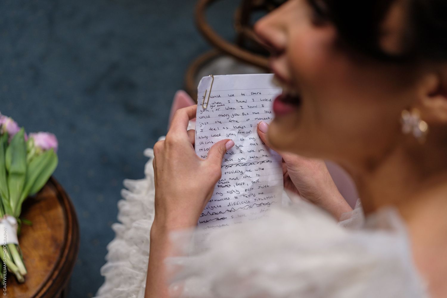 Bride in white dress reading handwritten wedding vows letter, holding paper with red lipstick and pearl earrings.