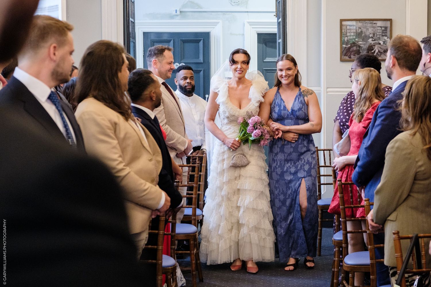 Bride in ruffled gown walks down aisle with bridesmaid in blue dress at elegant indoor wedding ceremony.