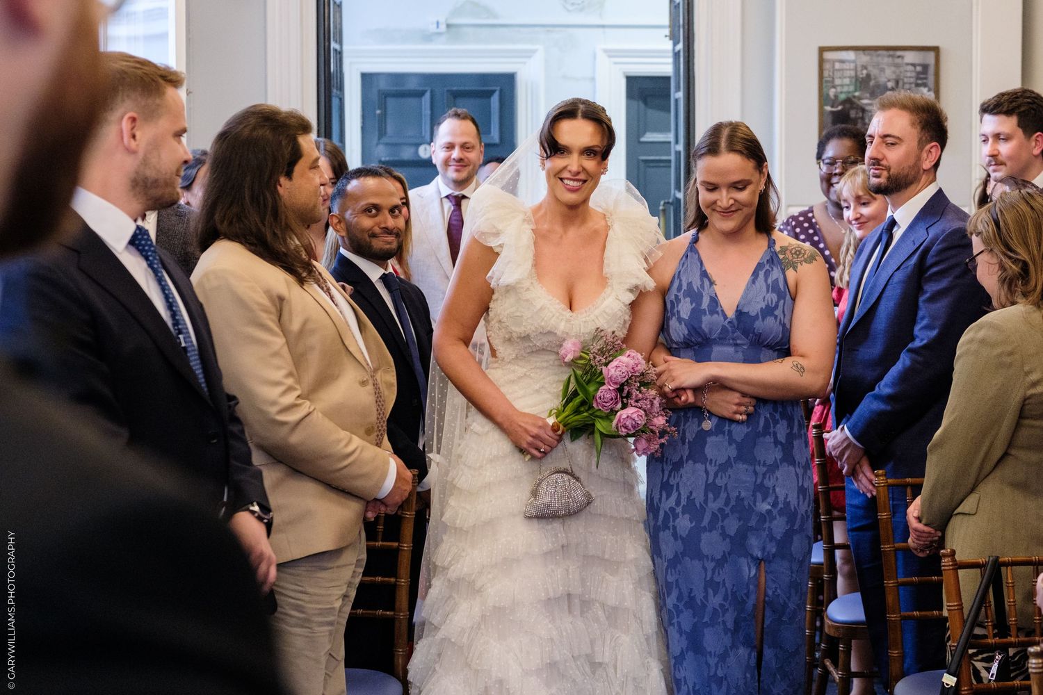 Bride walks down the aisle smiling, holding a bouquet, surrounded by wedding guests in a bright ceremony venue.