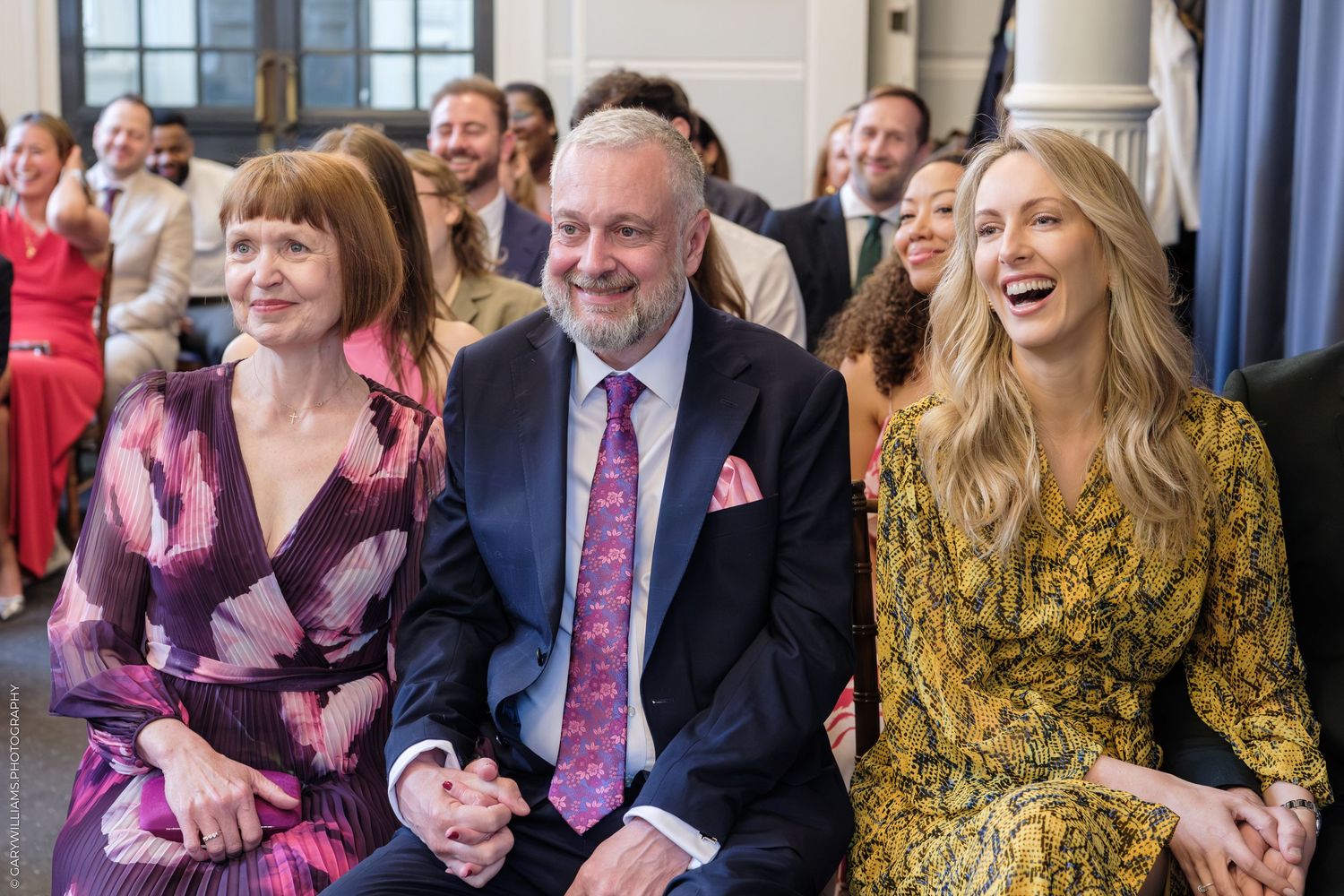 Three elegantly dressed guests laughing and smiling while seated at a formal indoor celebration or wedding ceremony.