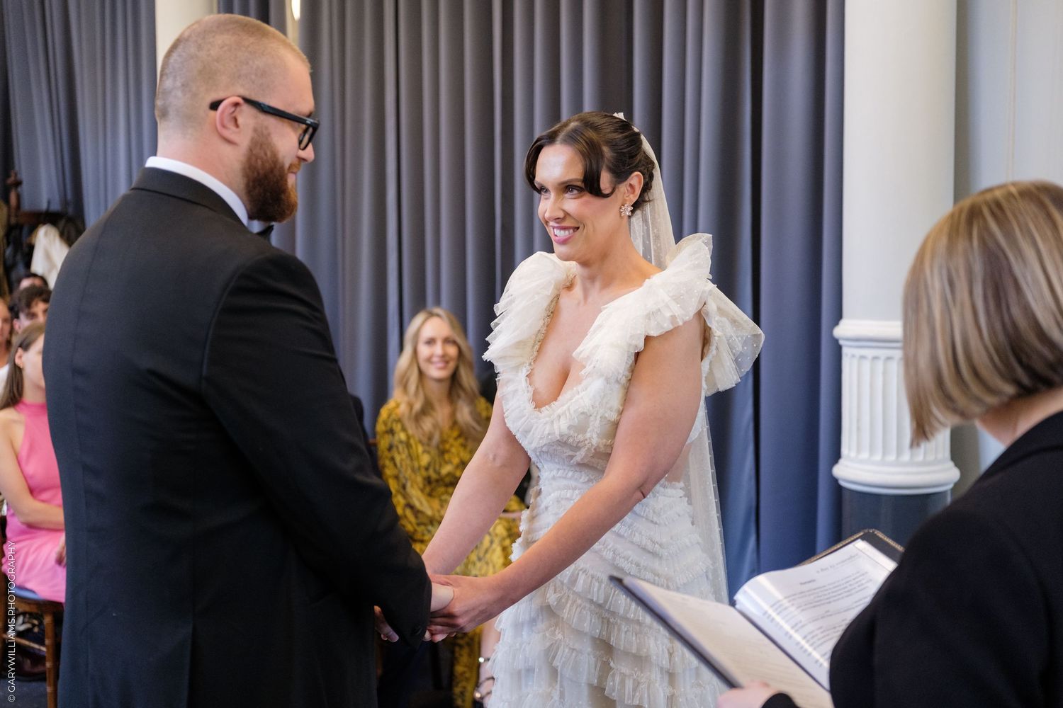 Bride and groom exchange vows at wedding ceremony, bride wearing elegant white ruffled gown, guests seated in background.