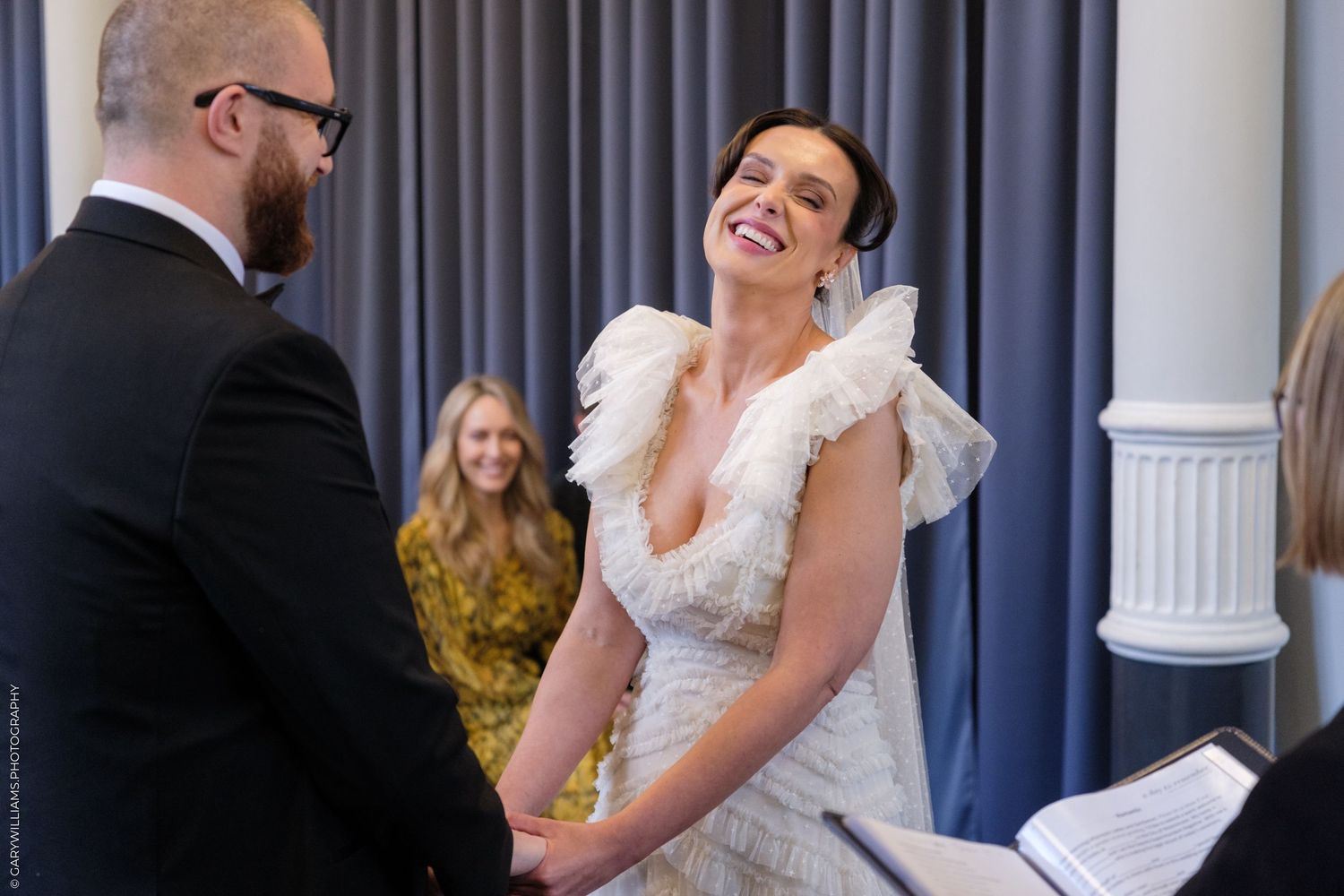 Bride in white feathered gown laughing joyfully while holding hands with groom during indoor wedding ceremony.