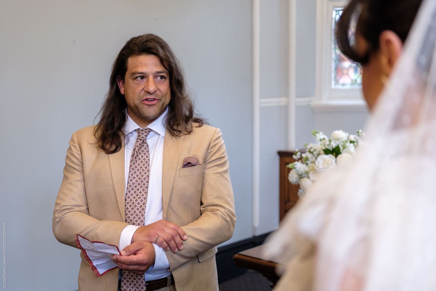 Groom in beige suit smiling at bride in white veil during wedding ceremony in elegant white chapel.