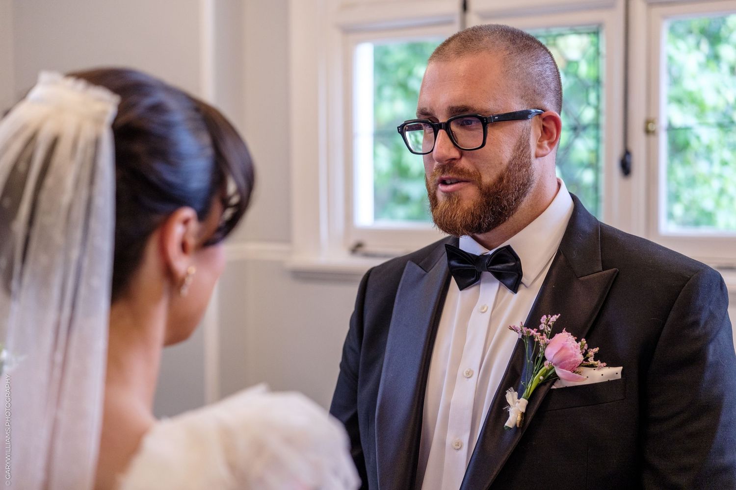 Groom in black tuxedo with bow tie and boutonniere exchanges vows with bride in white dress and veil.