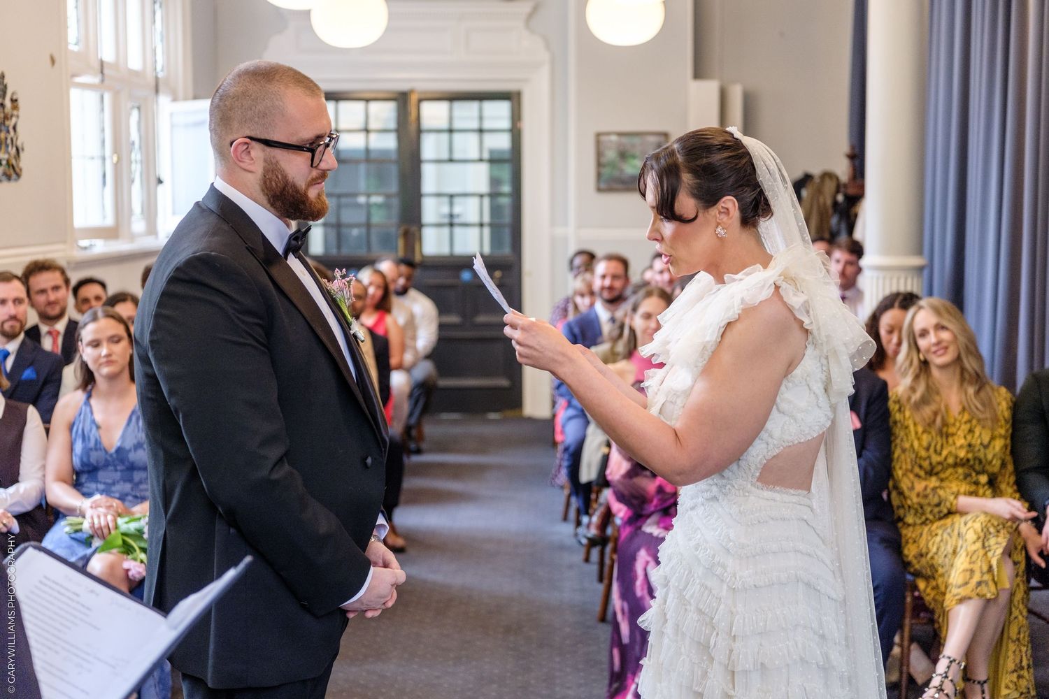Bride reads vows to groom in dark suit during intimate wedding ceremony with seated guests in background.