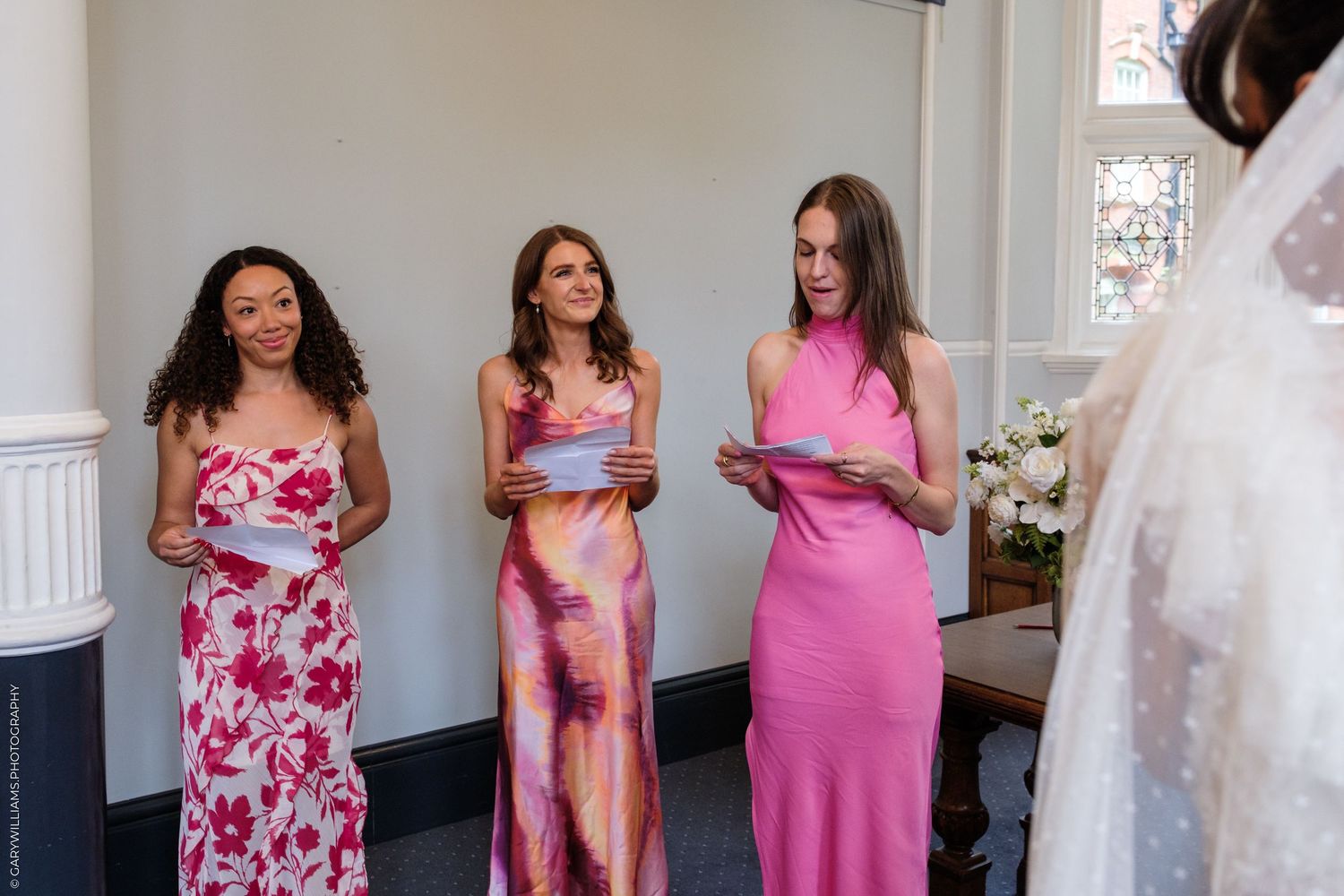 Three bridesmaids in pink dresses holding cards stand before a bride in a white wedding gown during a ceremony.