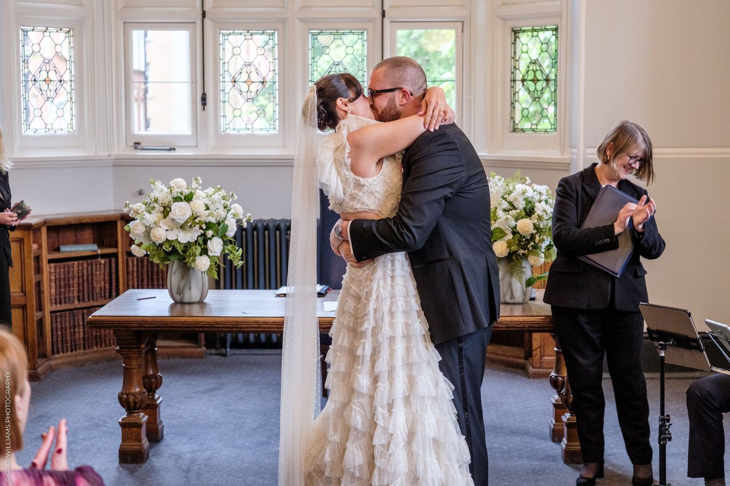 Bride and groom share first kiss at elegant indoor wedding ceremony with white floral arrangements and stained glass windows.