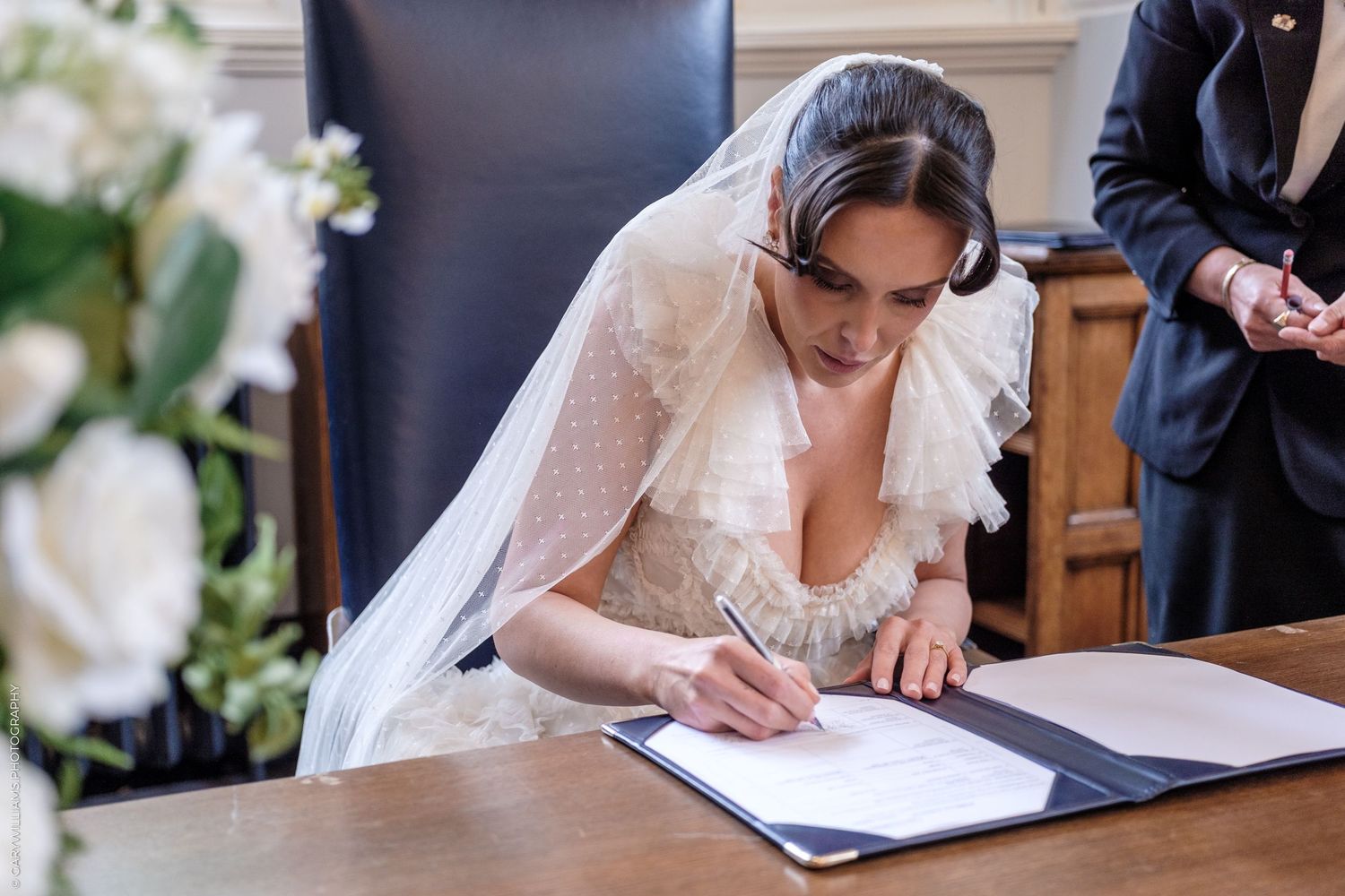 Bride in lace wedding dress and veil signing marriage register at wooden table during wedding ceremony.