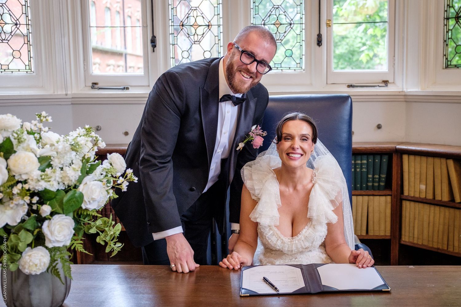 Bride and groom smiling at wedding registry signing table with white floral arrangement in elegant room.