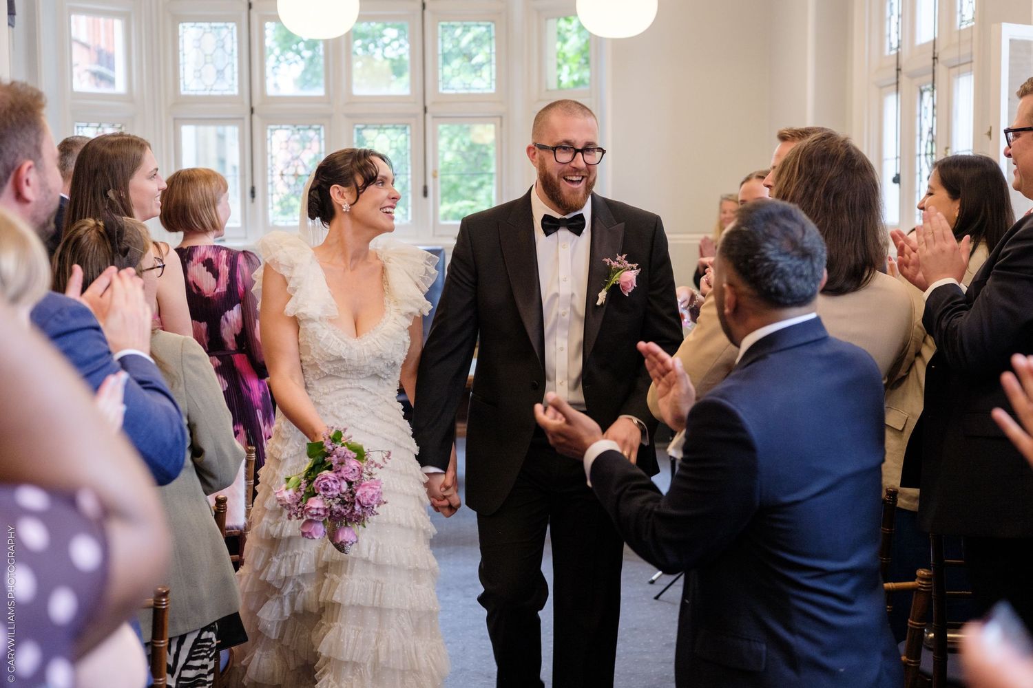 Bride and groom walk down the aisle smiling, holding hands, as wedding guests applaud in a bright ceremony hall.