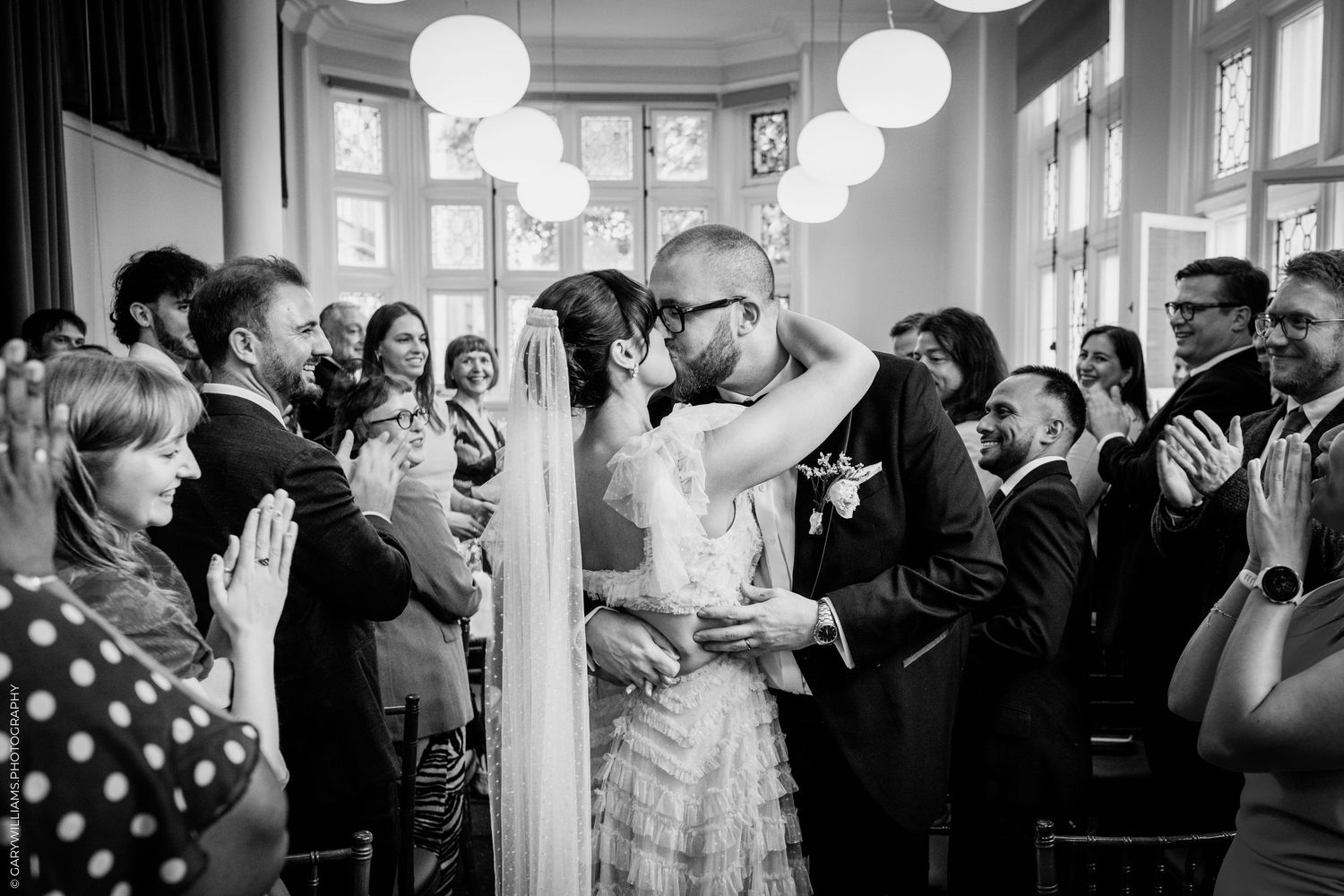 Black and white photo of a couple sharing their first kiss at a wedding ceremony surrounded by applauding guests.