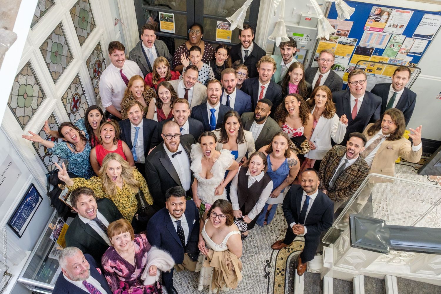 Large group of elegantly dressed wedding guests posing together for a photo from above in a bright venue.
