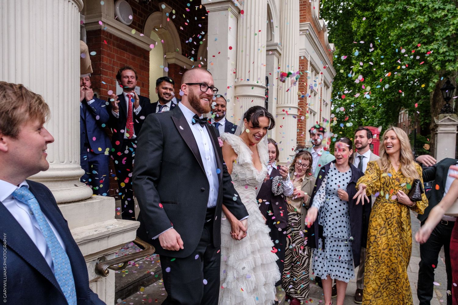 Joyful newlyweds exit church as guests throw colorful confetti during outdoor wedding celebration.