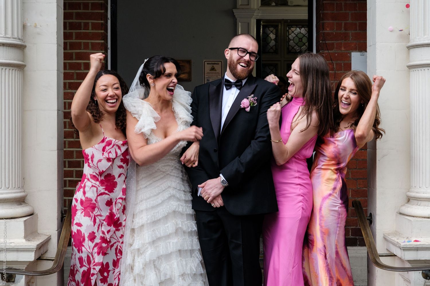 Joyful wedding party with bride in white ruffled gown, groom in tuxedo, and three bridesmaids in colorful dresses celebrating outdoors.