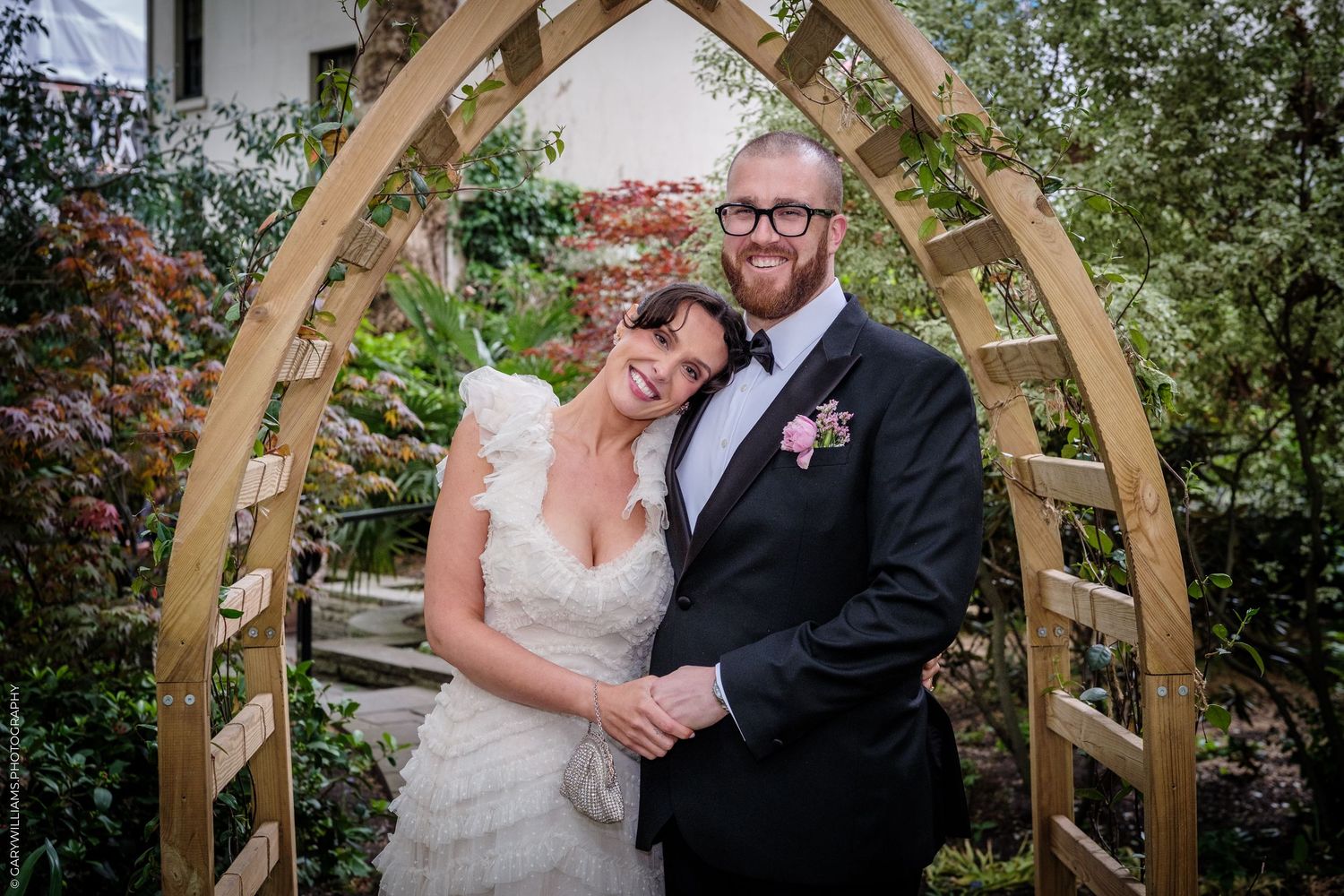 Bride and groom posing under a wooden arch in a lush garden, smiling on their wedding day.