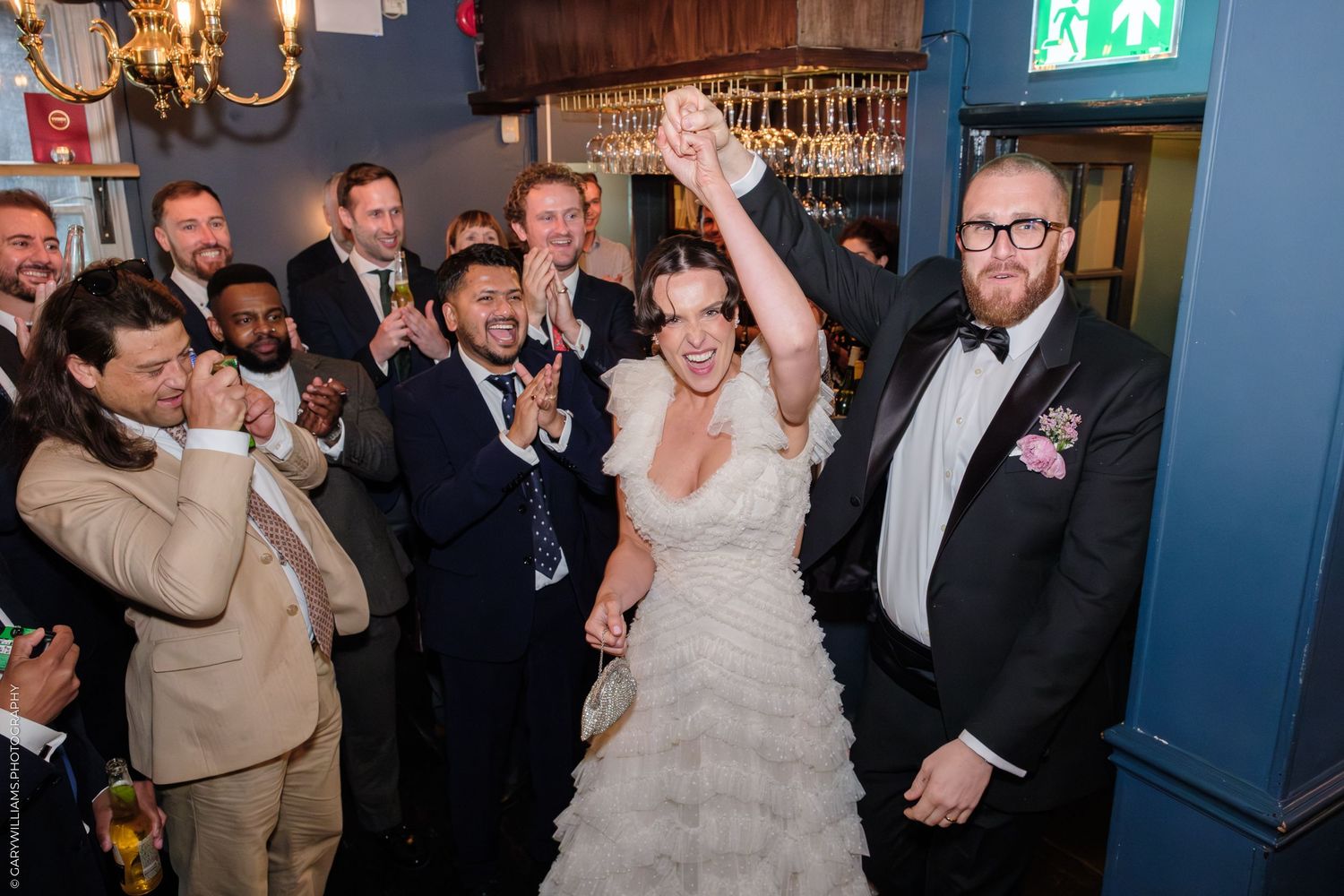 Bride celebrates joyfully with raised arms surrounded by cheering guests in suits at a wedding reception.
