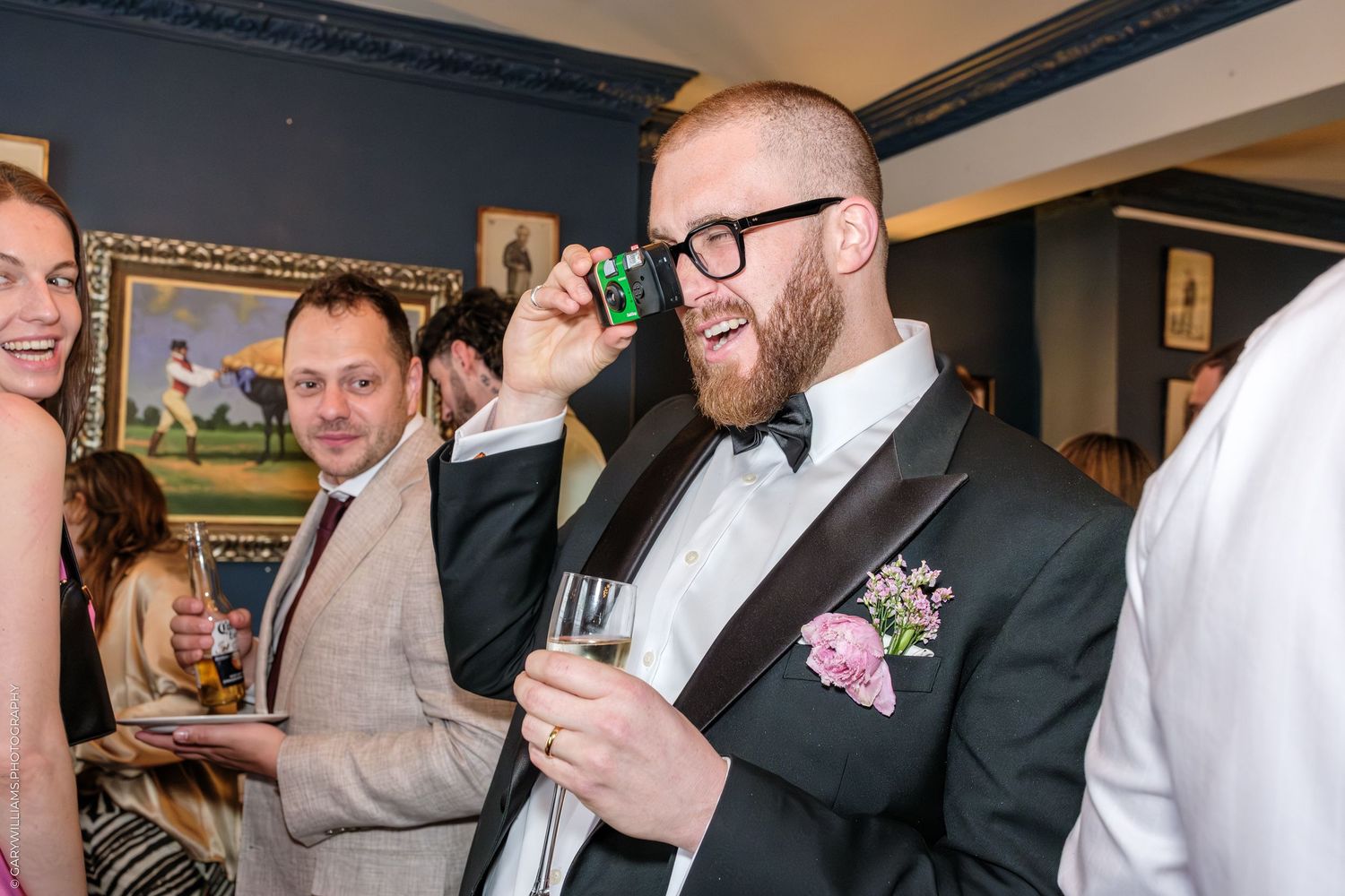 Man in tuxedo with bow tie and boutonniere holds camera to eye at formal event, champagne glass in hand.