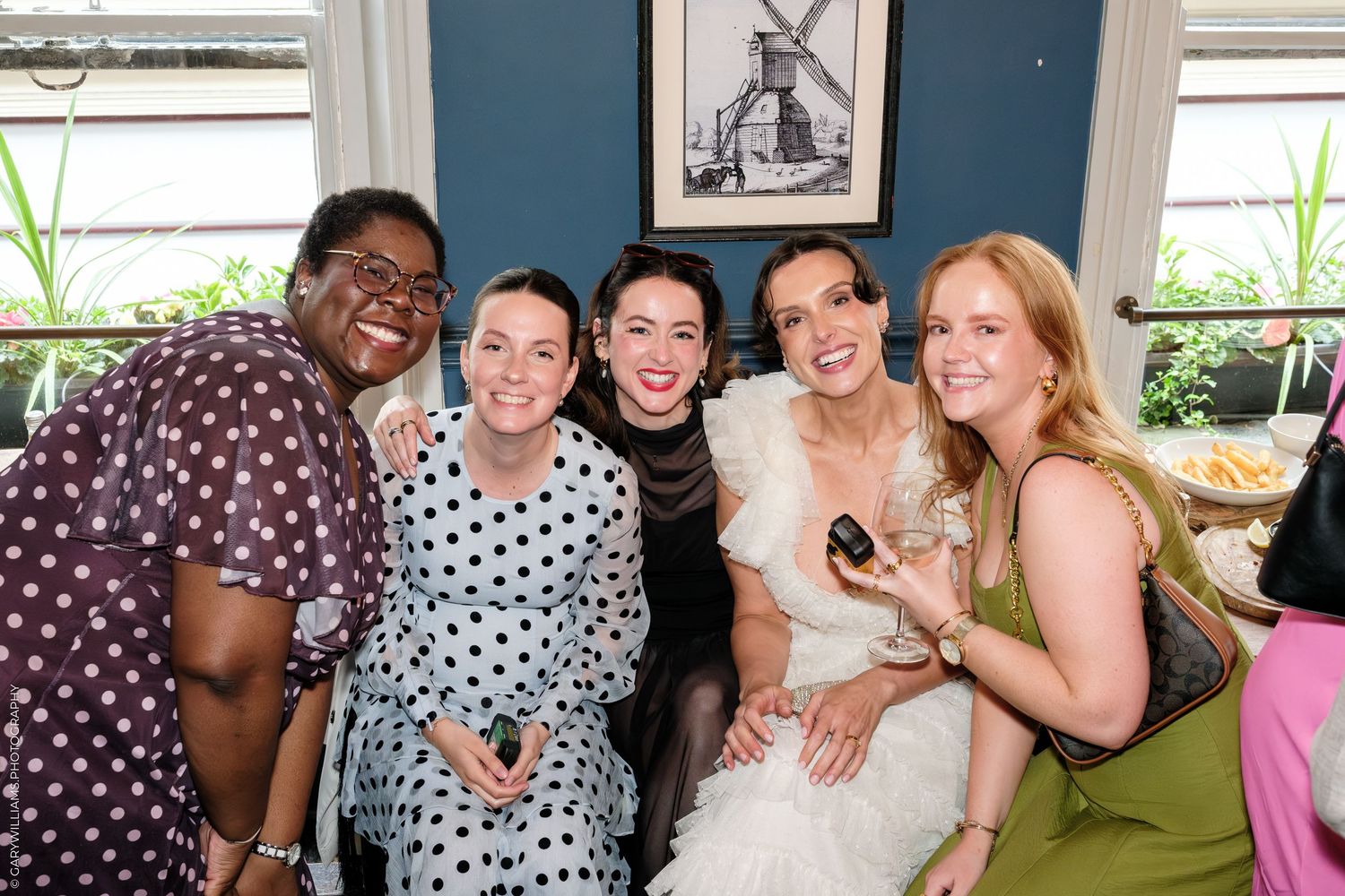 Five women smiling together at a wedding reception, bride in white dress surrounded by friends in colorful outfits.