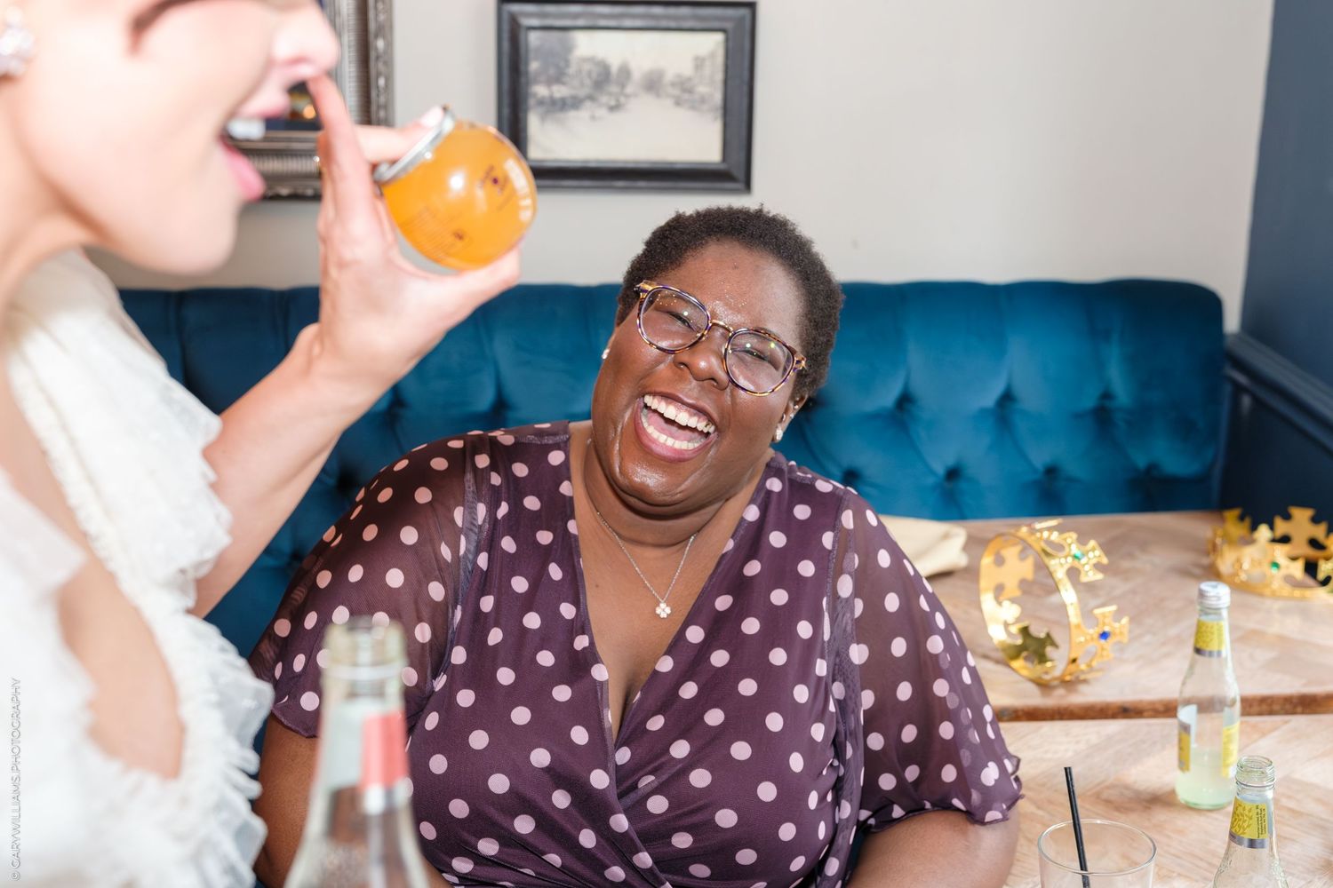 Two women laughing and enjoying drinks together on a blue velvet sofa in a cozy indoor setting.