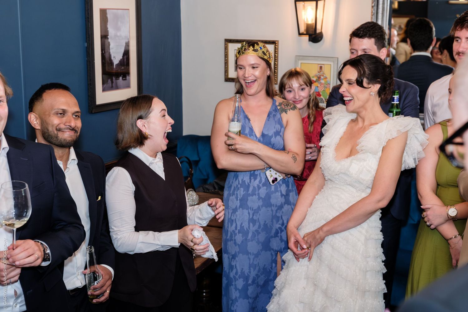 Bride in white gown laughing with guests at wedding reception in blue-walled venue.