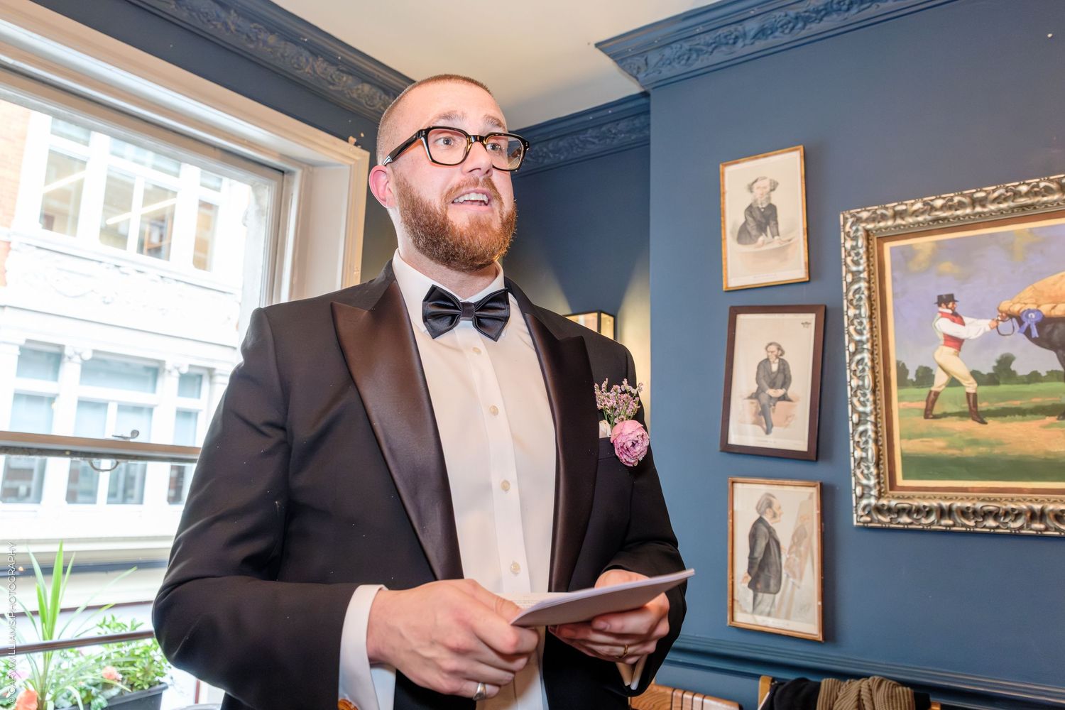 A smiling groom in a black tuxedo with bow tie and boutonniere holds a card near a window with framed art.