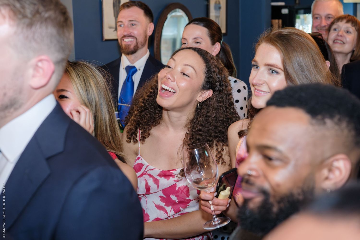 Group of smiling adults socializing at a party, woman in floral dress laughing with wine glass.