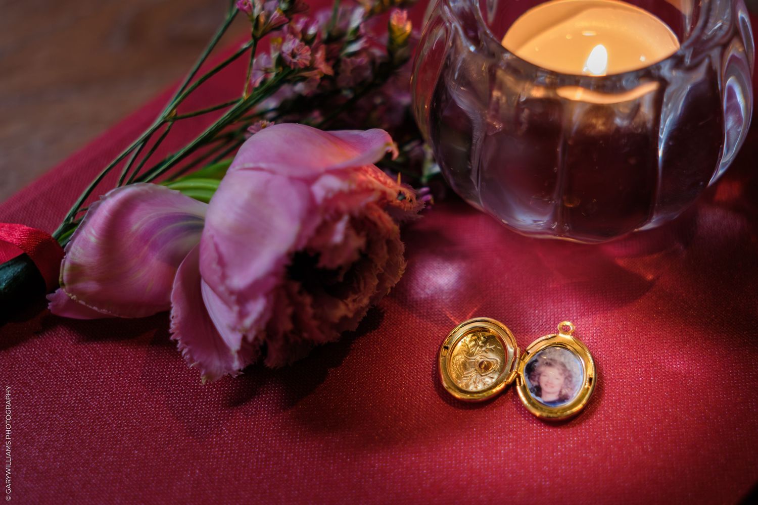Gold heart locket open with photo beside pink tulip, candle, and flowers on red satin surface.