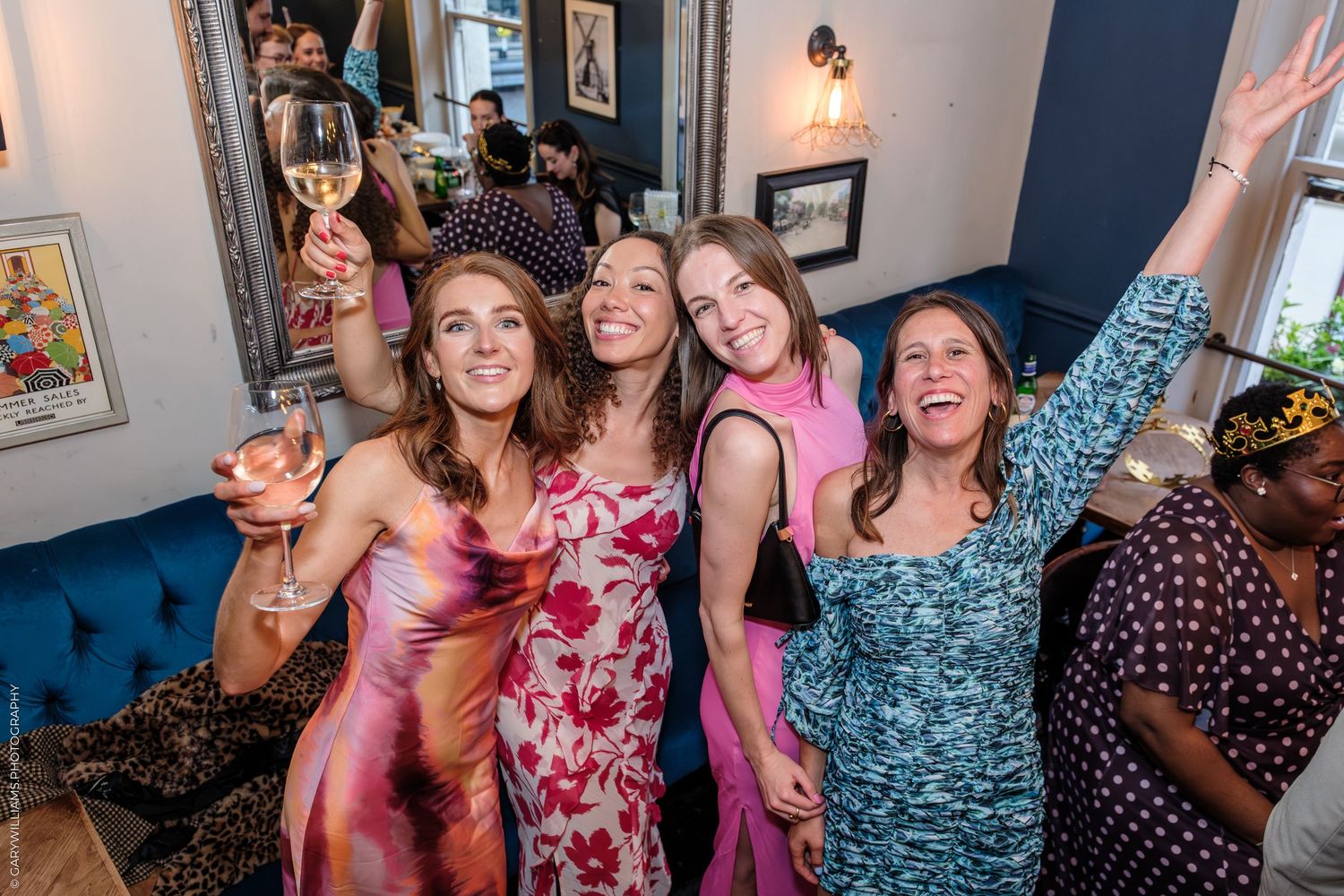 Four women smiling and celebrating at a party, holding wine glasses and raising arms in a festive indoor setting.