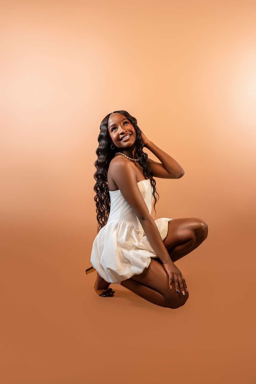 Smiling Black woman in white strapless mini dress crouching on beige background with long curly hair.