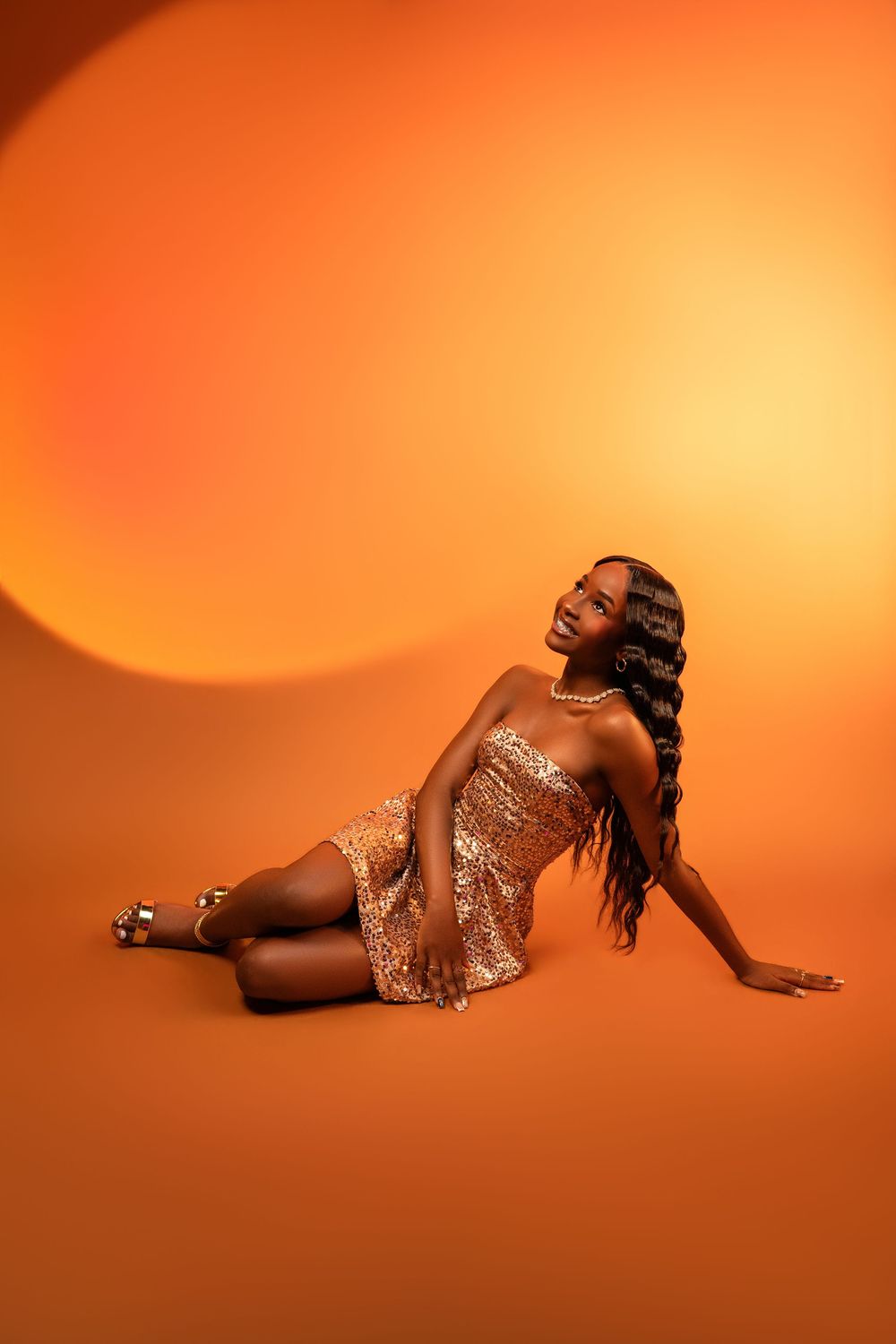 A Black woman in a gold sequin dress sits gracefully on an orange background, smiling upward with long wavy hair.