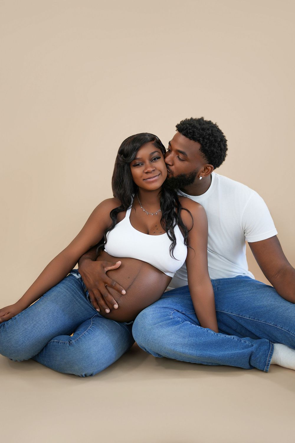 A pregnant Black woman in white crop top and jeans sits with her partner who kisses her cheek on a beige background.