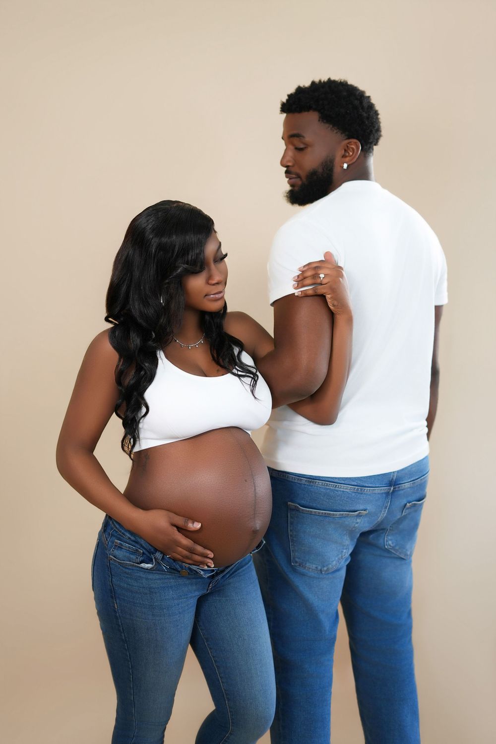 Pregnant Black couple in white tops and jeans posing together, woman cradling baby bump, against beige background.