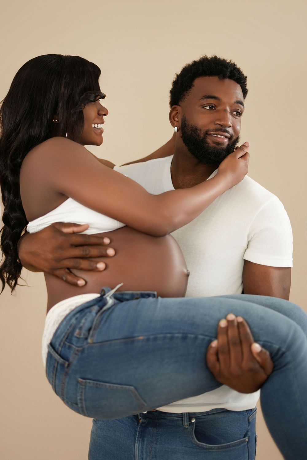 A man carries a pregnant woman in his arms as she smiles and touches his face, both wearing white tops and jeans.