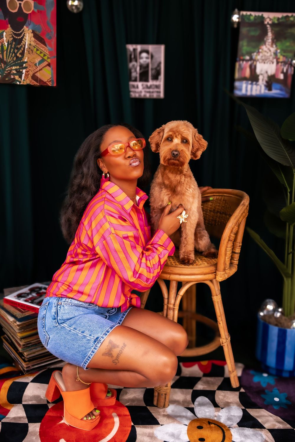 Woman in pink striped shirt and denim shorts poses with a golden doodle dog on a rattan chair in a colorful room.