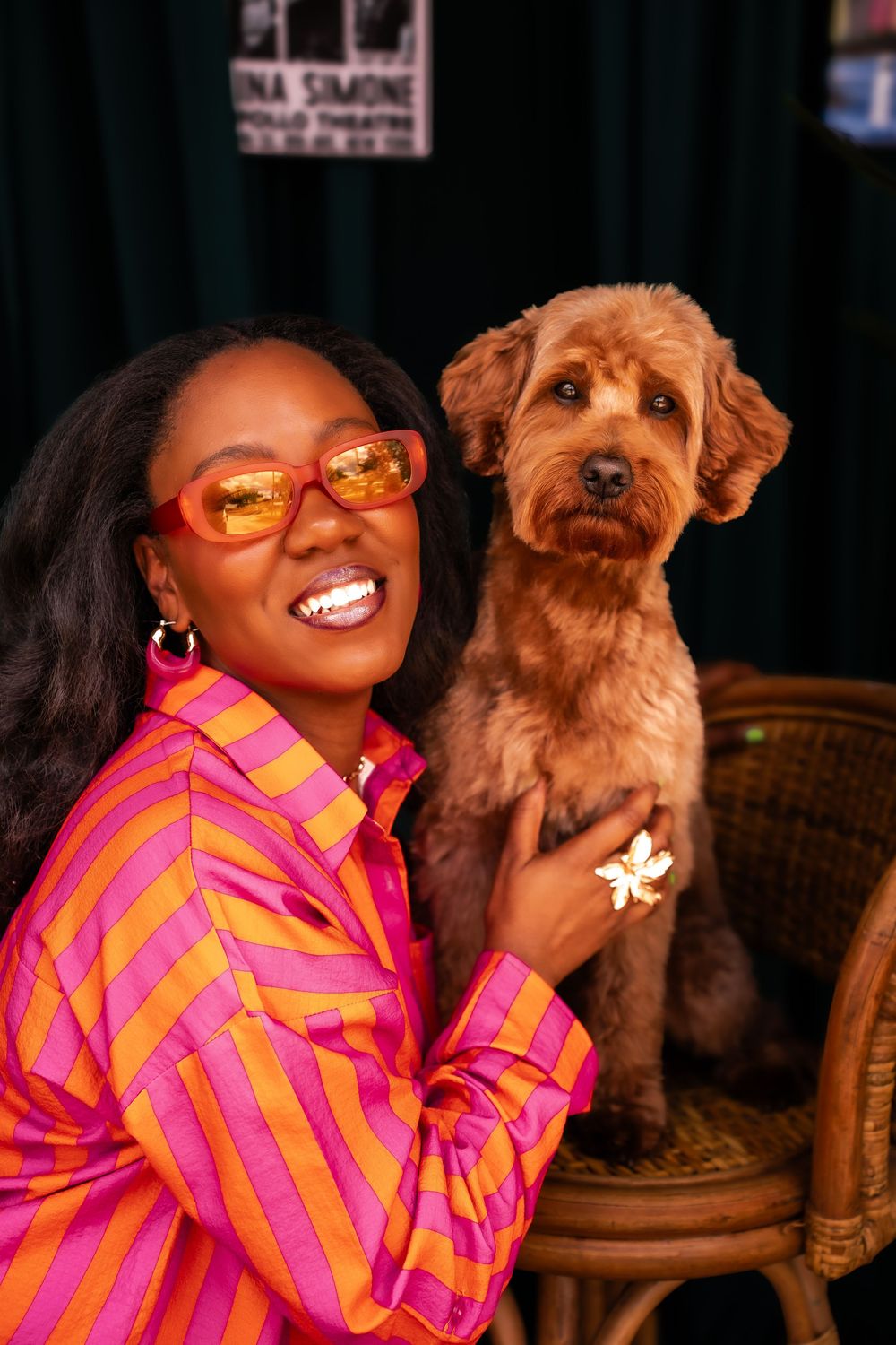Smiling woman in colorful striped shirt and orange sunglasses poses with a fluffy golden doodle dog on a wicker chair.